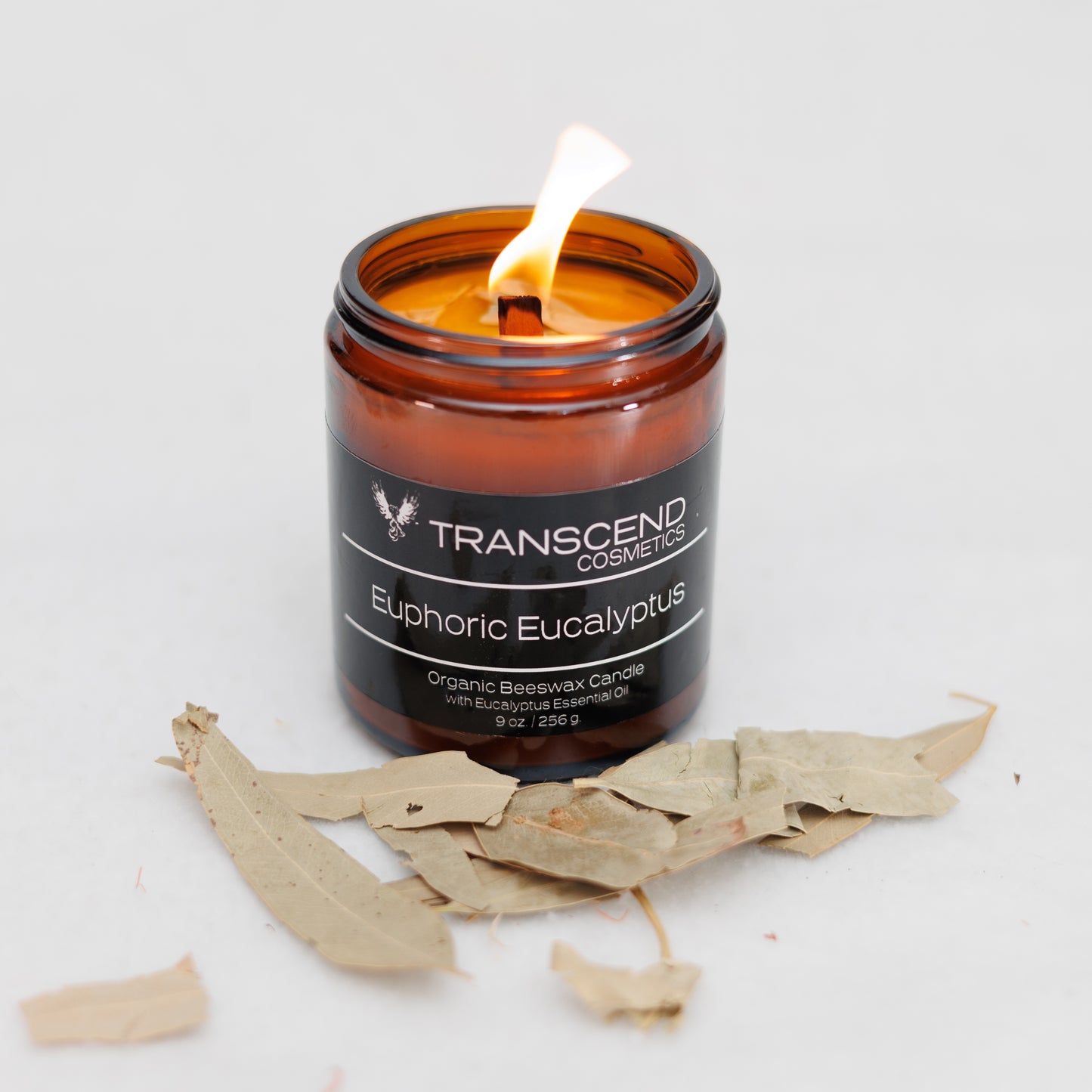 Transcend Cosmetics Euphoric Eucalyptus Organic Beeswax Candle in an amber glass jar with a wooden wick, burning with a warm flame, surrounded by dried Eucalyptus leaves, highlighting its natural and soothing ingredients.
