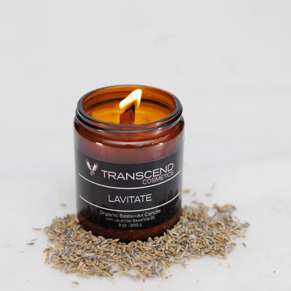 Transcend Cosmetics Lavitate Organic Beeswax Candle in an amber glass jar with a wooden wick, burning with a warm flame, surrounded by dried Lavender petals, highlighting its natural and soothing ingredients.