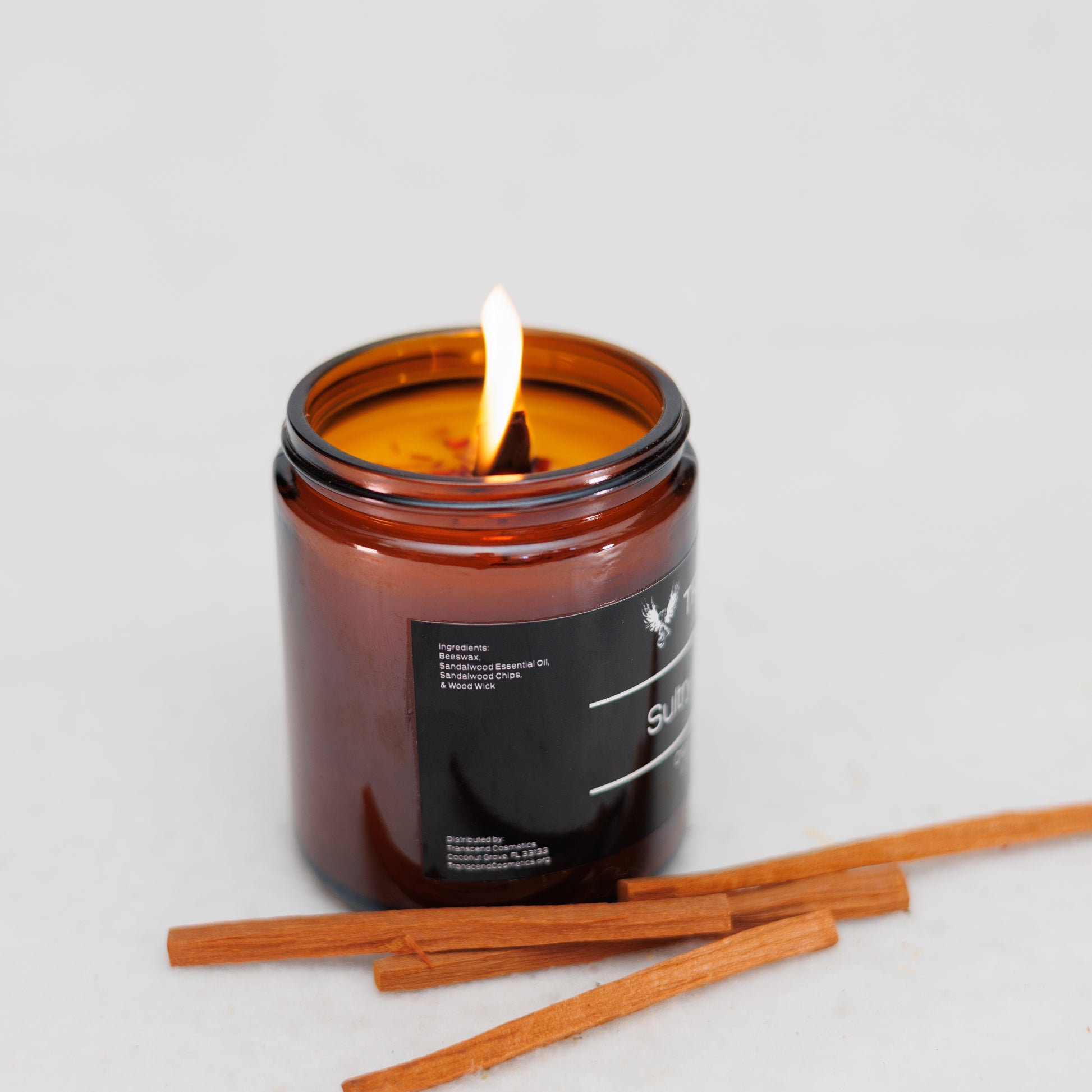 Image showing the ingredient list on the Transcend Cosmetics Sultry Sandalwood Organic Beeswax Candle, in an amber glass jar with a wooden wick, burning with a warm flame, surrounded by Sandalwood sticks, highlighting its natural and soothing ingredients.
Ingredients are: Beeswax, Sandalwood Essential Oil, Sandalwood Sticks, & Wooden Wick. 