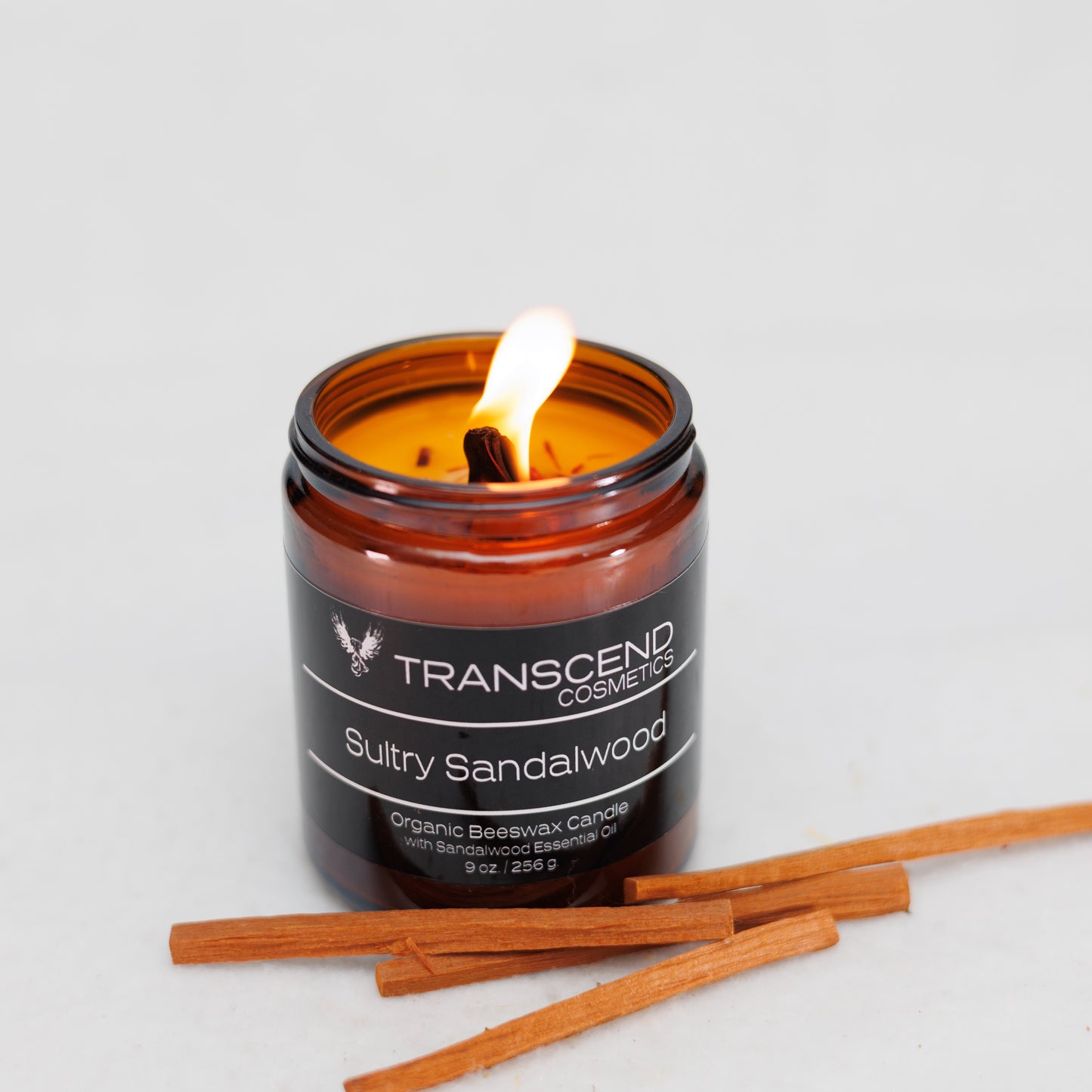 Transcend Cosmetics Sultry Sandalwood Organic Beeswax Candle in an amber glass jar with a wooden wick, burning with a warm flame, surrounded by Sandalwood sticks, highlighting its natural and soothing ingredients.